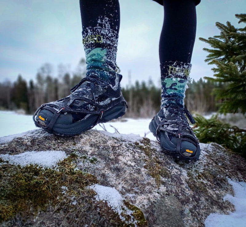 Load image into Gallery viewer, Person wearing snow boots and camo waterproof socks socks standing on a rock with a snowy landscape and trees in the background.