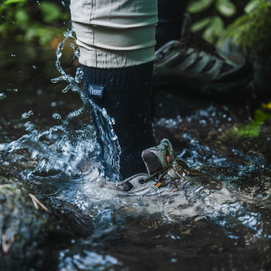A close-up action shot of a person's legs and feet wading through splashing water in a forest stream. The person is wearing light beige hiking pants, black waterproof socks, and gray hiking boots.