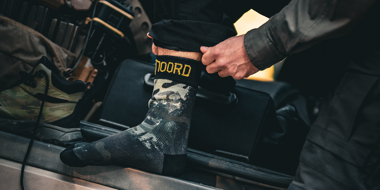 Military person putting on waterproof socks with camouflage pattern and 'Noord' branding, standing in the boot of a vehicle with tactical gear and hiking boots.