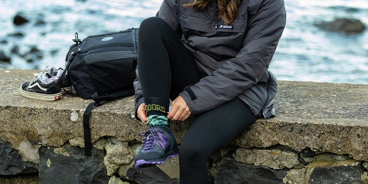 waterproof socks for women, sitting on a stone wall by the water wearing a gray jacket and black pants, with a backpack and shoes nearby.