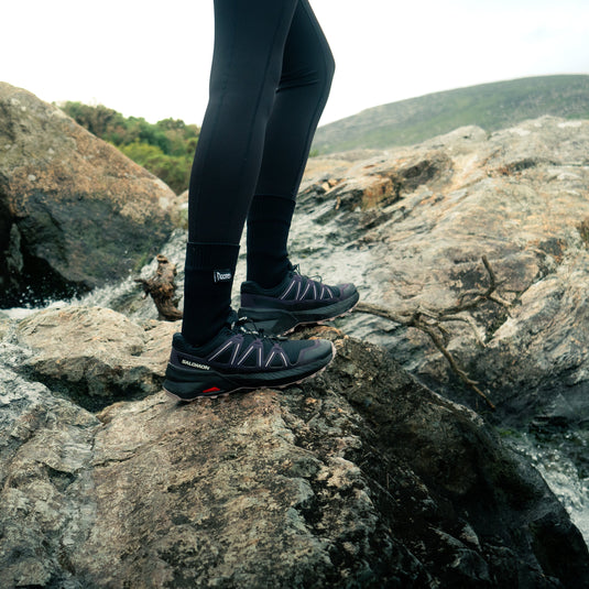 A person wearing black Salomon hiking shoes, noord waterproof socks and black leggings stands on large rocks outdoors, with water and a hazy landscape in the background.