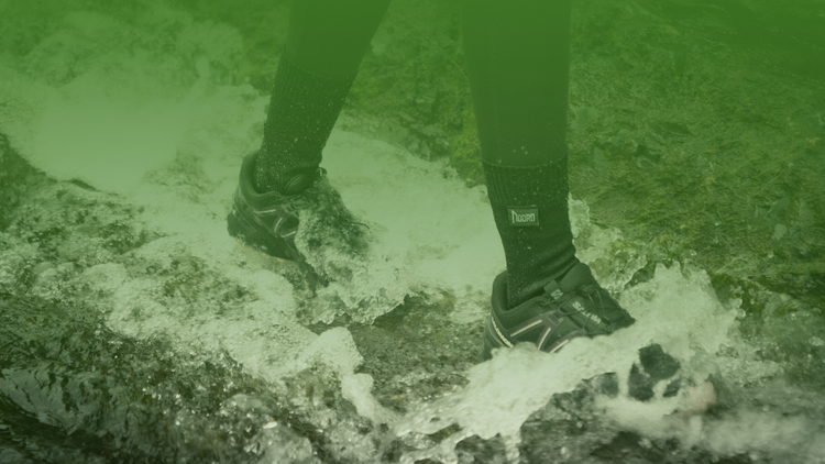 A person standing in shallow, foamy water wearing black athletic waterproof socks and black salomon trail-running shoes, with a strong green tint over the photo.
